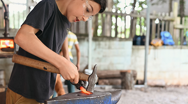 Blacksmithing student crafts a knife blade with hammer and anvil at summer camp