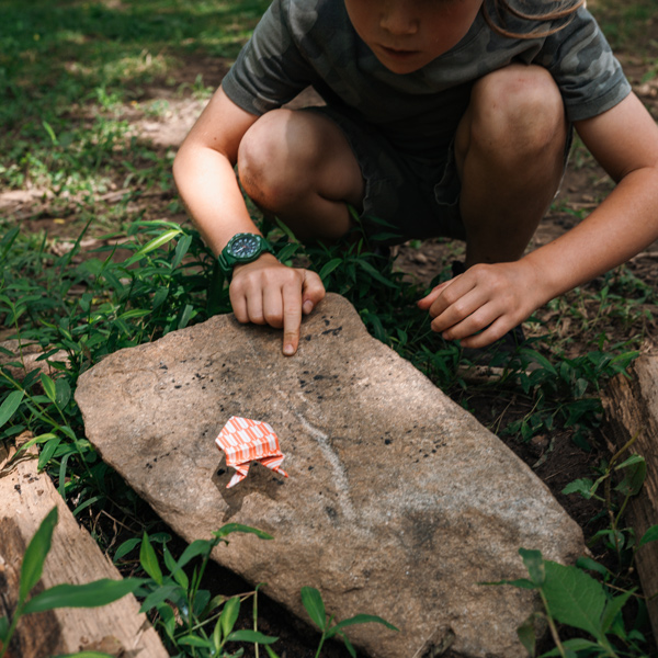 Boy crouching by a rock deep ijn the forest with an origami frog toy