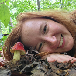 Naturalist and child mentor Breanne Jones close up head shot lying on forest floor next to a mushroom poking up thru leaf litter