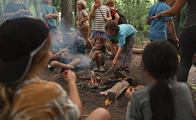 Children gather around a campfire at forest's edge in Asheville at a day program during Hurricane Helene recovery days