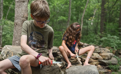 Children carving tools from branches seated atop boulders in the woods at Asheville forest homeschool day