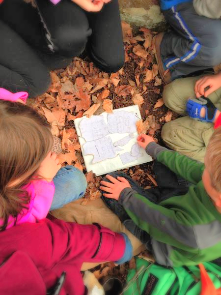 Children gathered in a circle around a journal sitting on the forest floor at camp