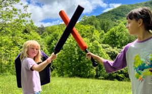 Day campers square off with foam boffing swords at the start of play in a field along the woods