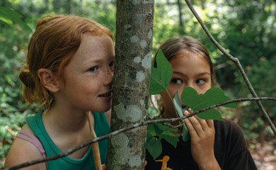 Two girls hiding behind a trea looking like they are mischevious and having fun in the forest at camp